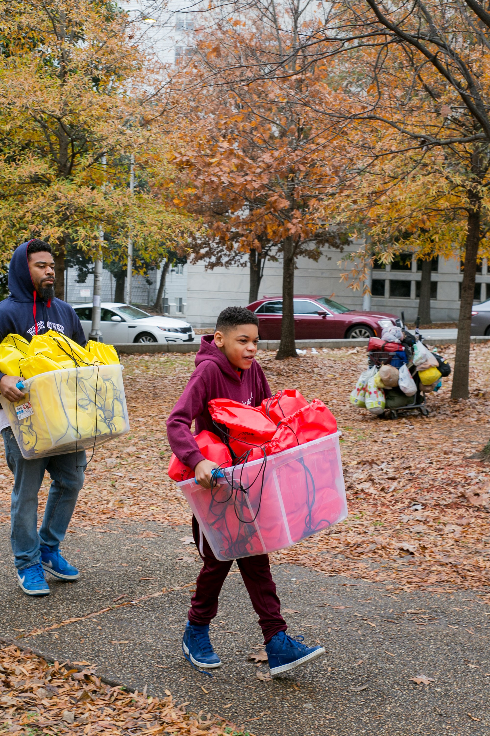 A man and a boy carrying bins of yellow and red care bags down a leaf-covered path