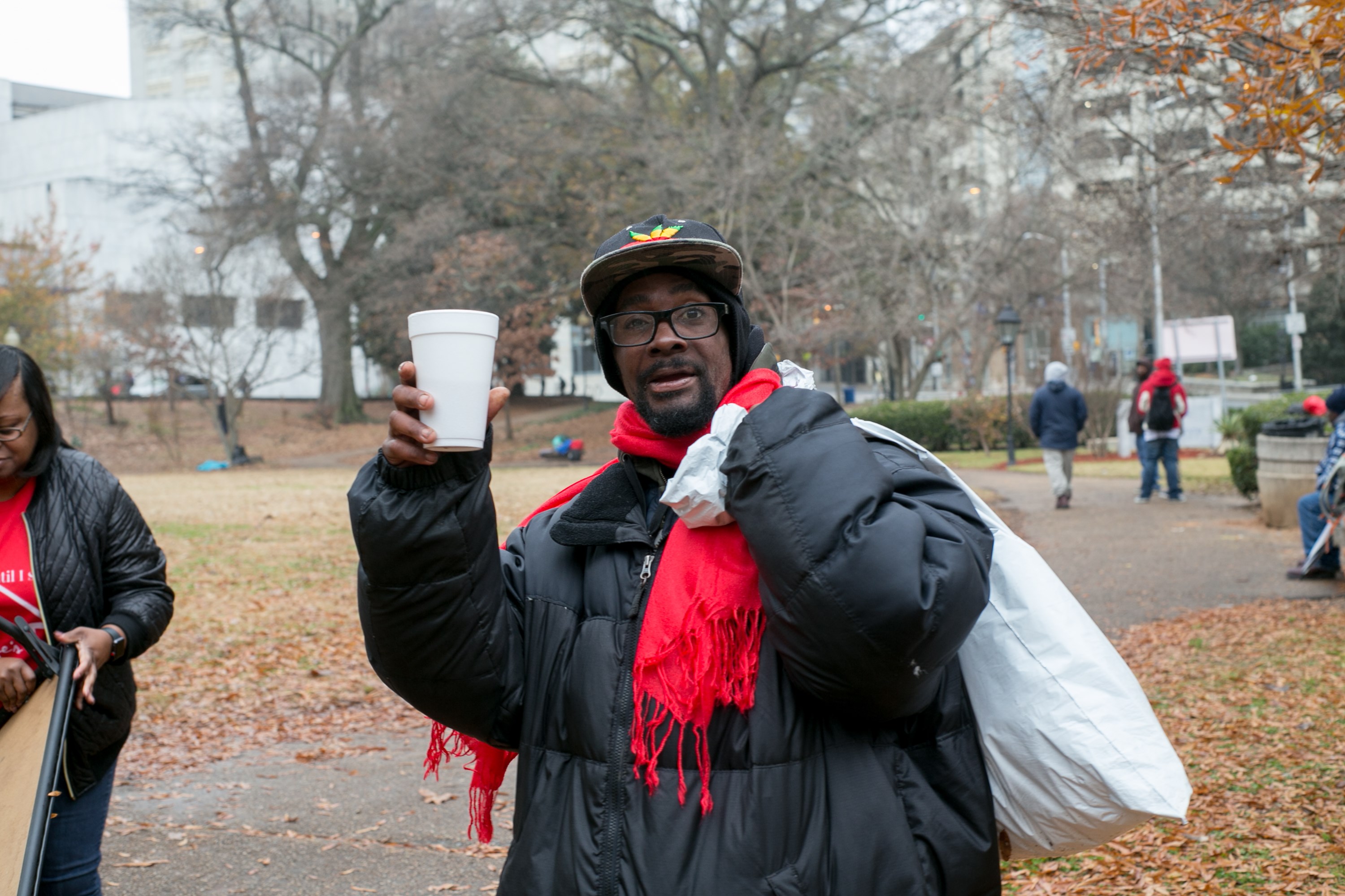 A man in a red scarf holding a warm drink and a bag of supplies, smiling