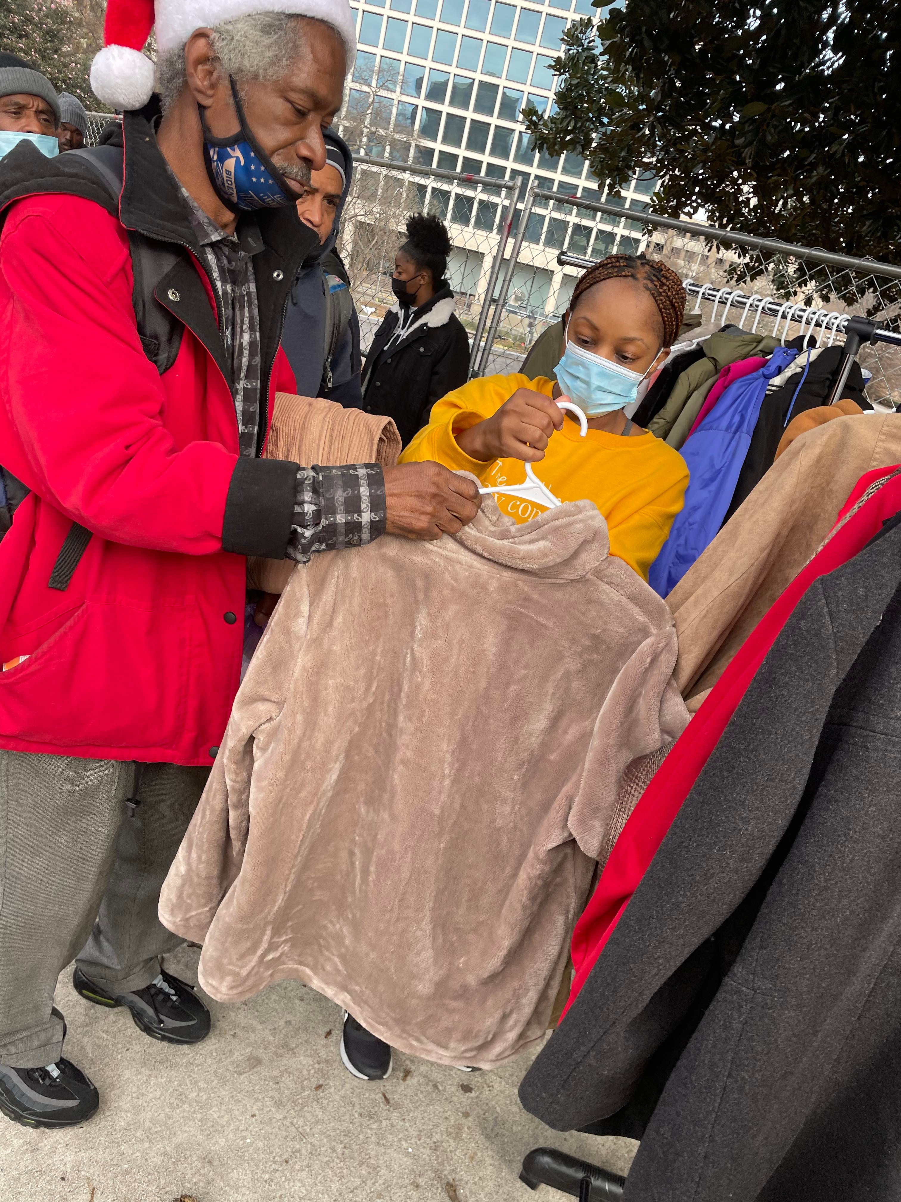 A volunteer helping a man in a Santa hat pick out a warm coat