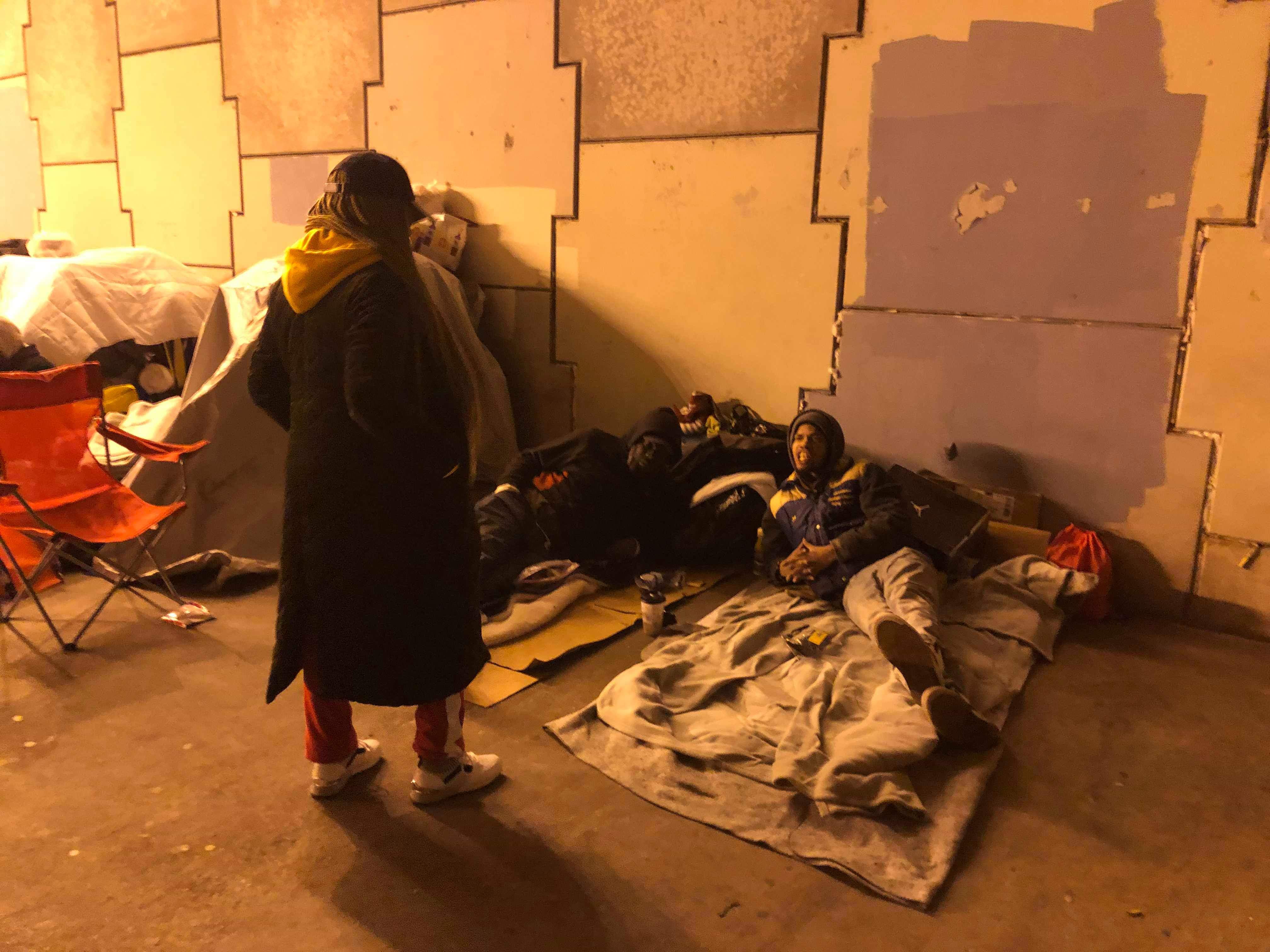 A volunteer talking with two men resting under blankets against the underpass wall