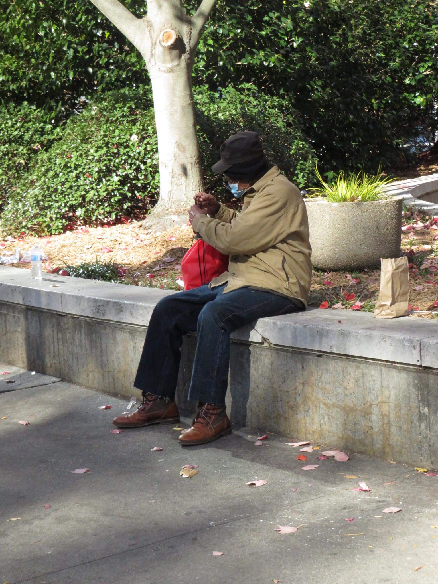 A man in a tan jacket and cap sitting on a concrete ledge with a red Jo-Anne's House care bag beside him