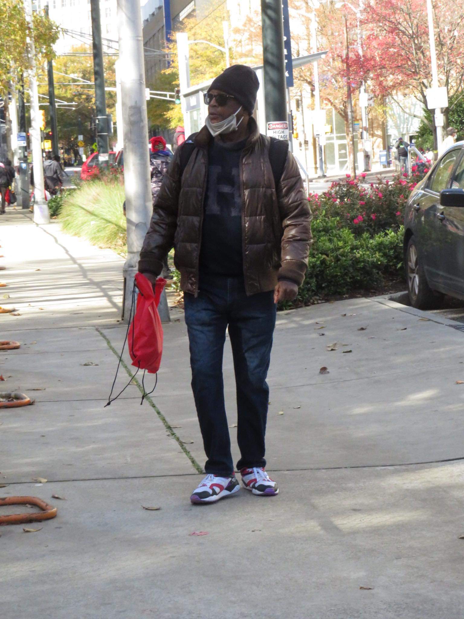 A man in a brown puffer jacket walking down the sidewalk carrying a red Jo-Anne's House care bag