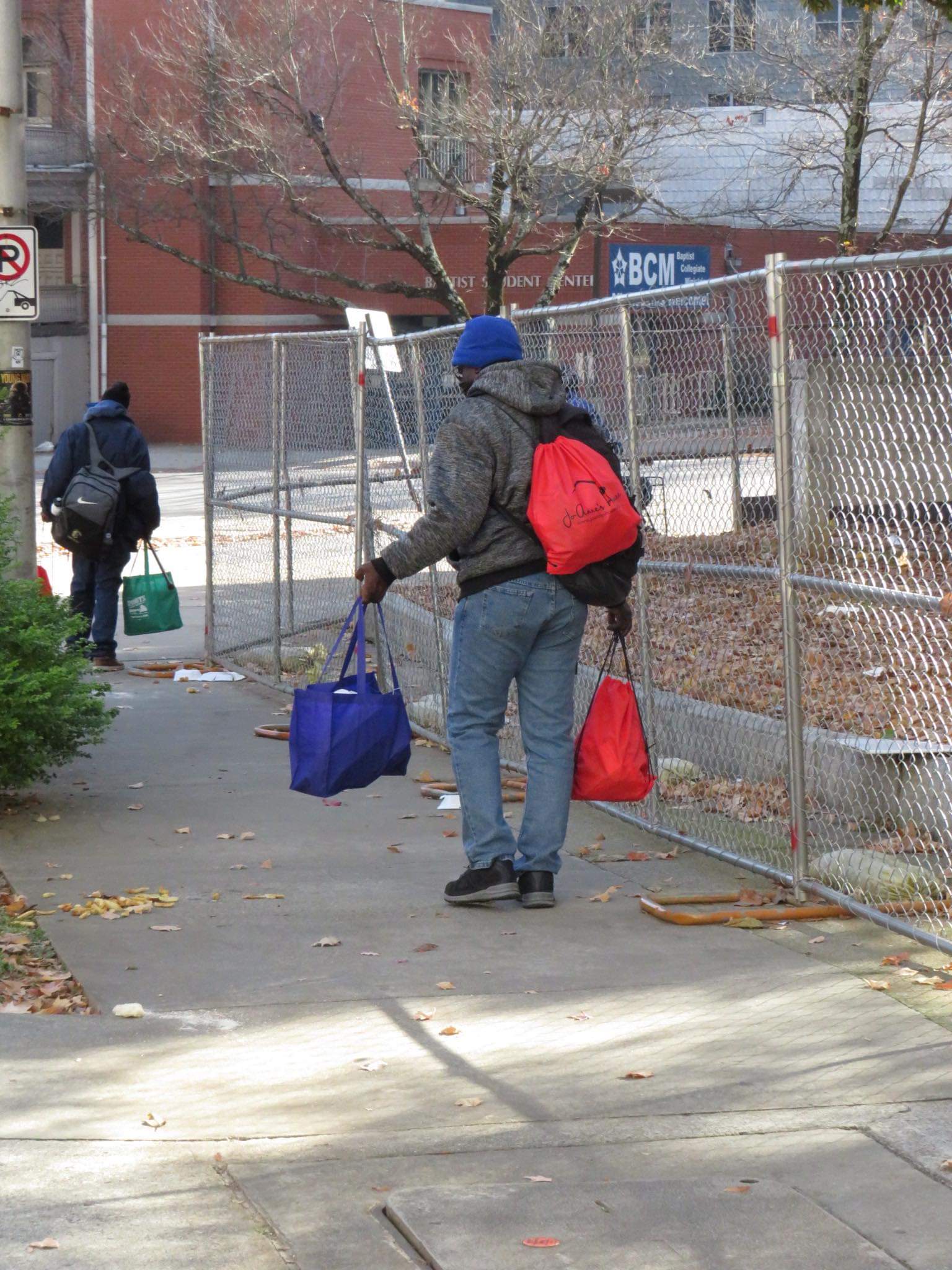 Two men walking away carrying red Jo-Anne's House care bags near a campus building